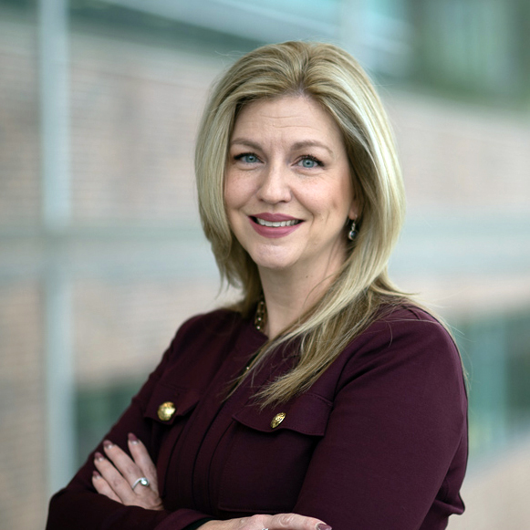 Dr. Kelly is wearing a claret coloured top and is standing in front of windows in the medical school atrium.