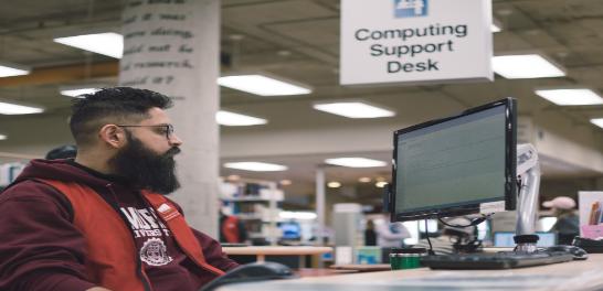 A Commons student staff member (wearing a red vest) works on a computer at the Computing Support Desk