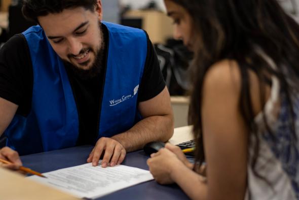 A smiling Writing Centre tutor (wearing a blue staff vest) assists a student with her paper.