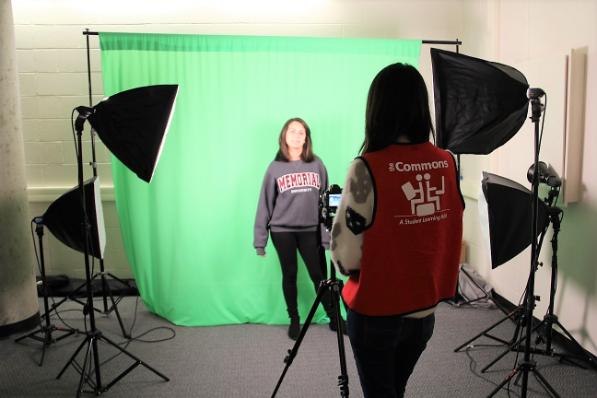 A student stands in front of a greenscreen background. A Commons employee (wearing a red staff vest) takes her pictures with a DSLR camera.
