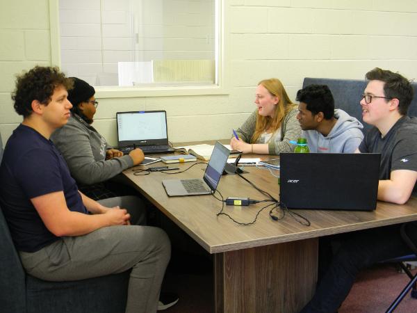 5 students sit around a collaborative workstation. They are working on 3 laptops and have notebooks in front of them.