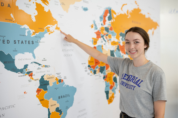 A student wearing a Memorial University t-shirt smiles while pointing to Newfoundland and Labrador on a large, colorful world map displayed on the wall.